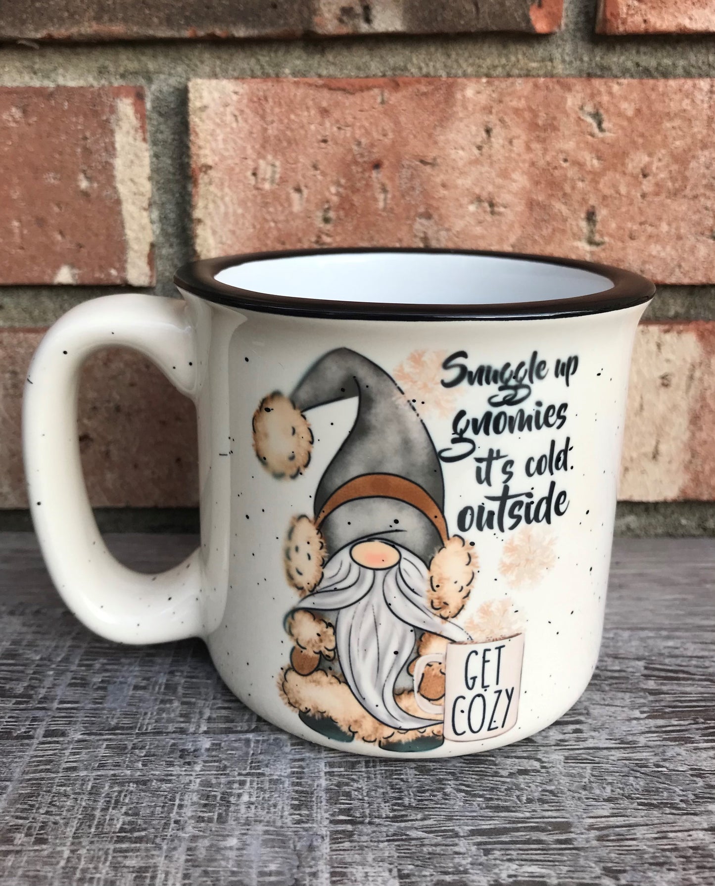 Camp style beige mug with speckled pattern that has a gnome on the front with a coffee cup that reads Snuggle up gnomies it’s cold outside