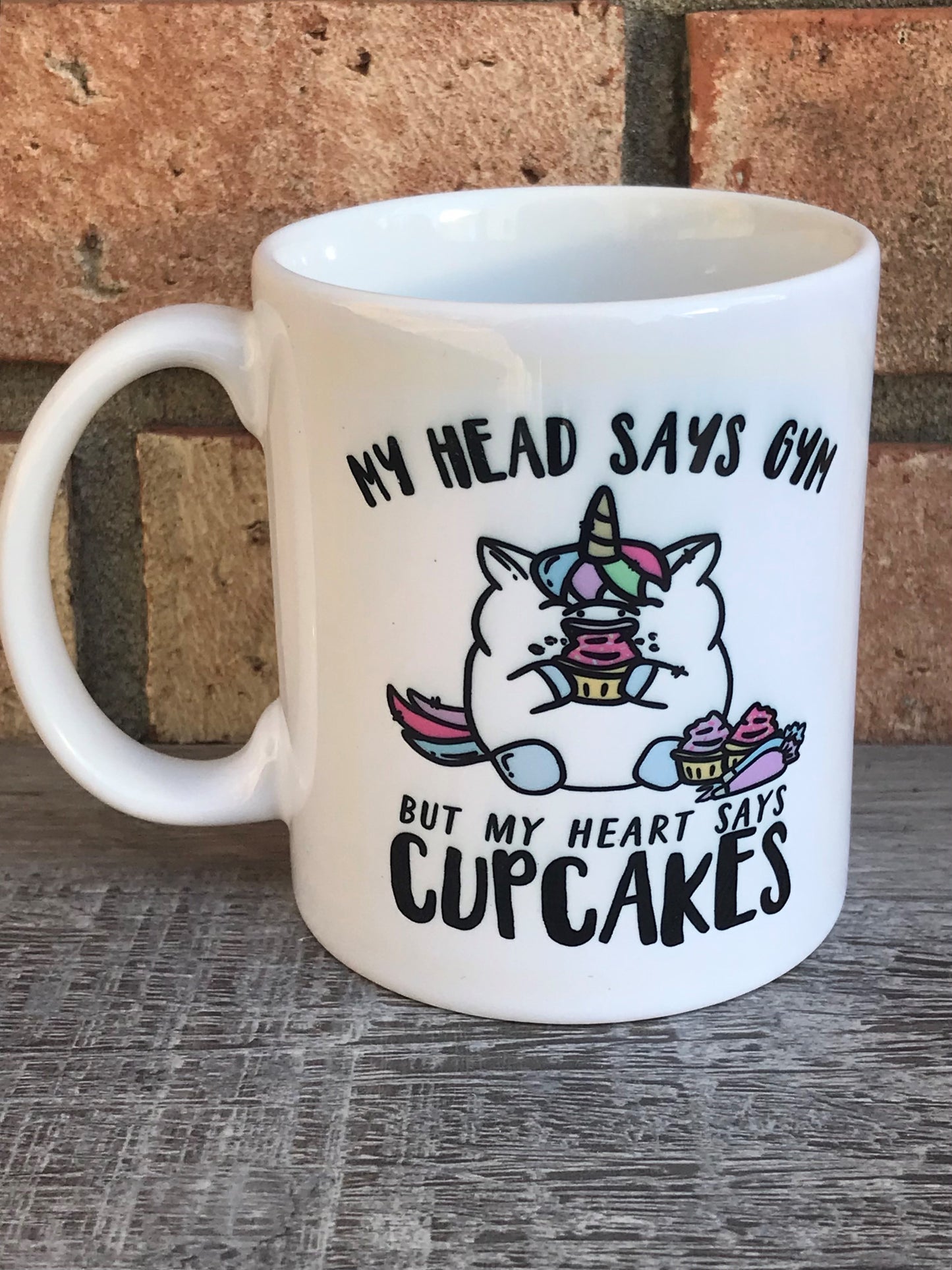 A white ceramic mug with a unicorn and text graphics related to cupcakes. mug says My head says gym but my heart says cupcakes