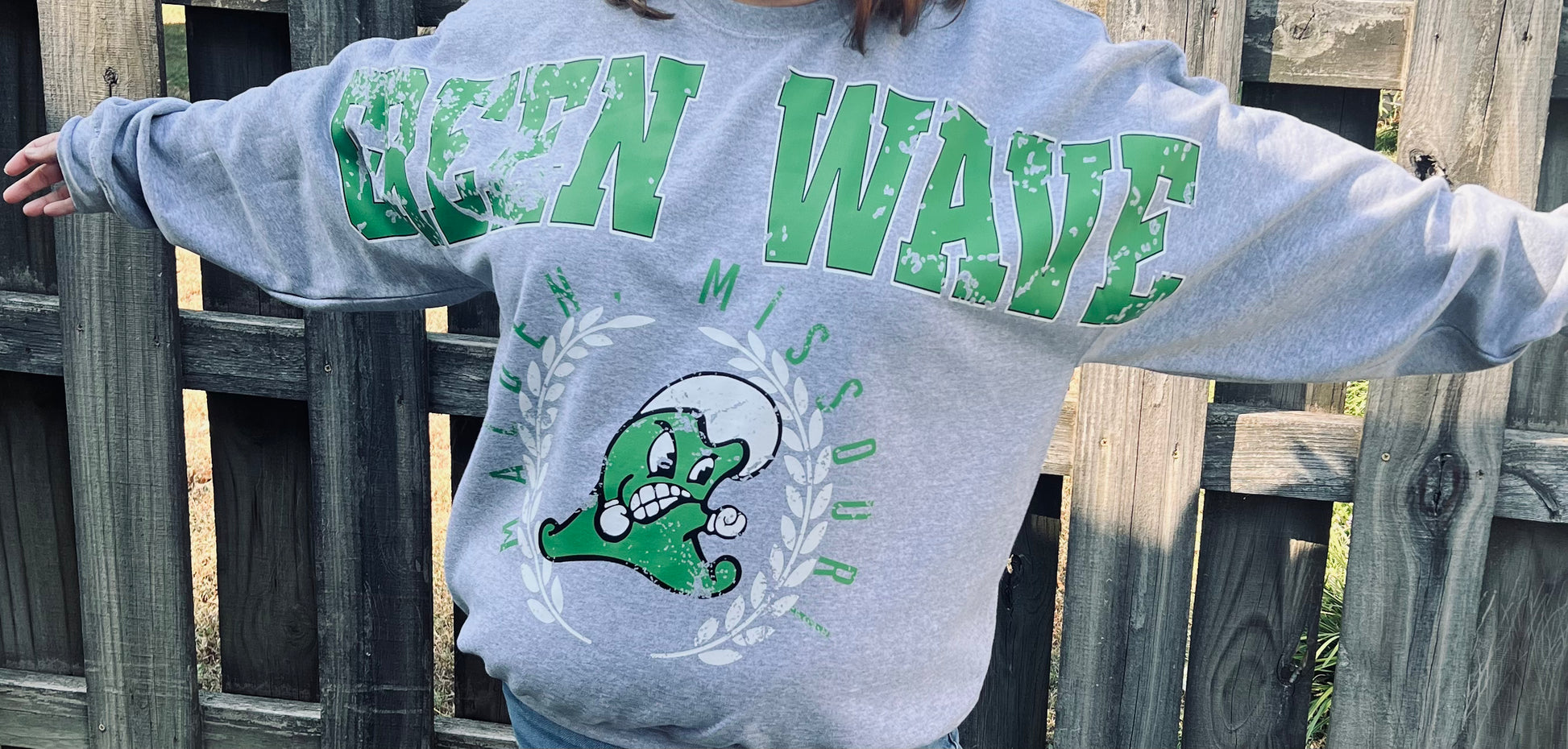 Person wearing a gray sweatshirt with green text and graphics, standing against a wooden fence.