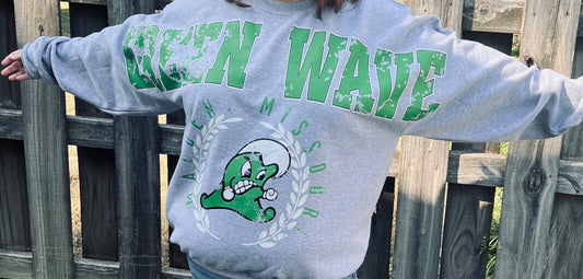 Person wearing a gray sweatshirt with green text and graphics, standing against a wooden fence.