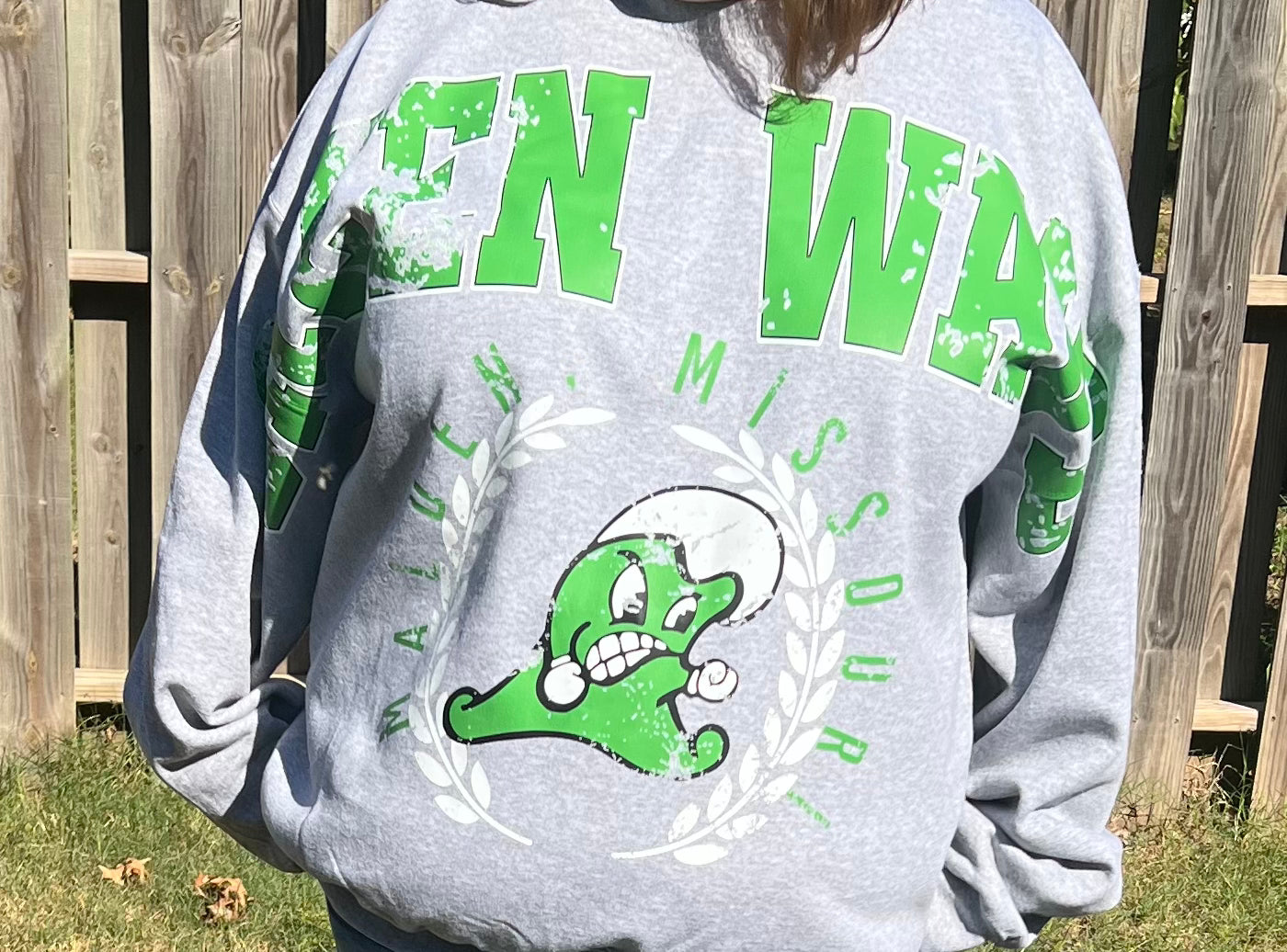 Person wearing a gray sweatshirt with green and white text and graphics, standing outdoors.