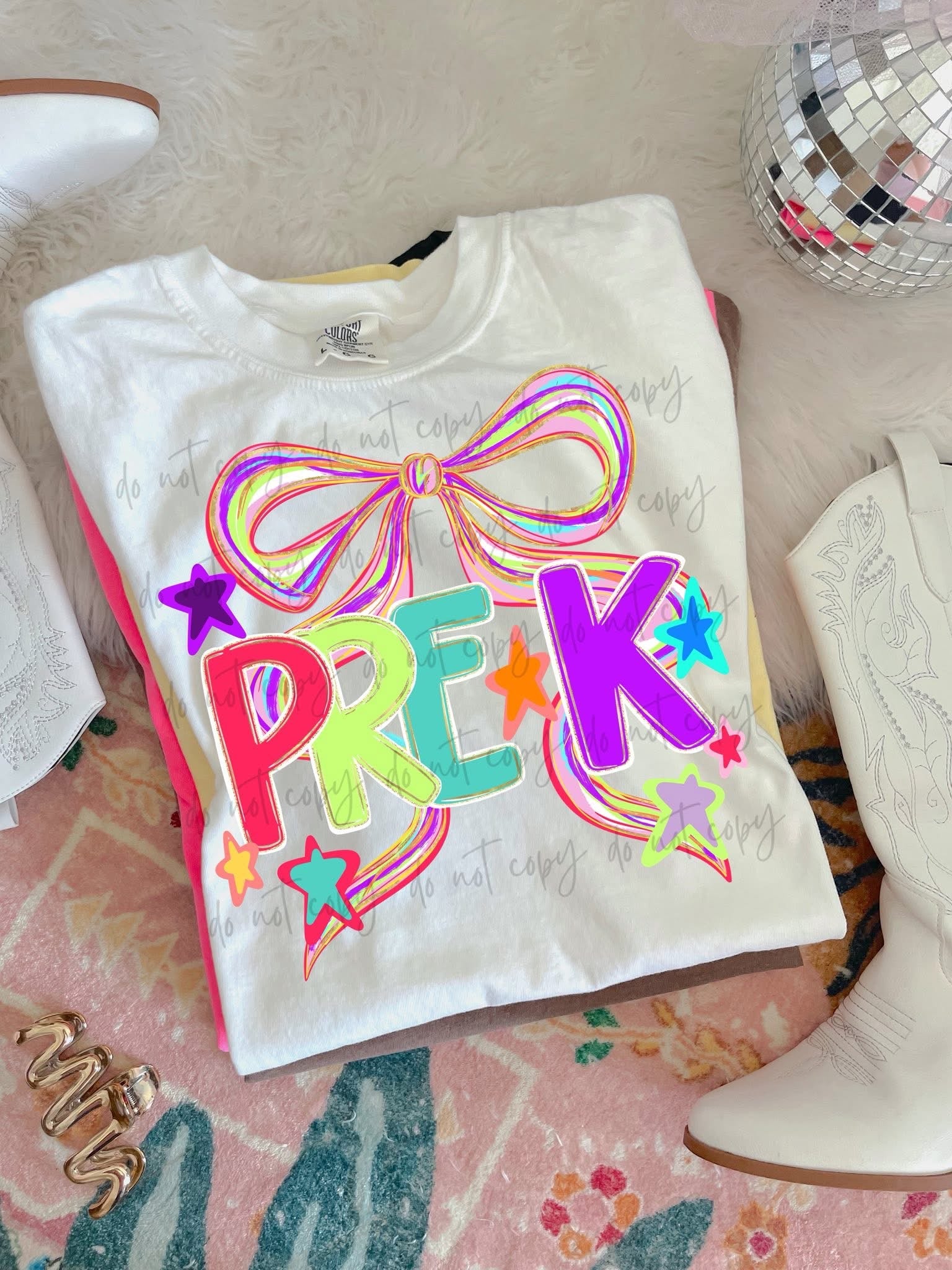 White t-shirt with colorful 'PREK' design on a pink surface with white boots and a disco ball.