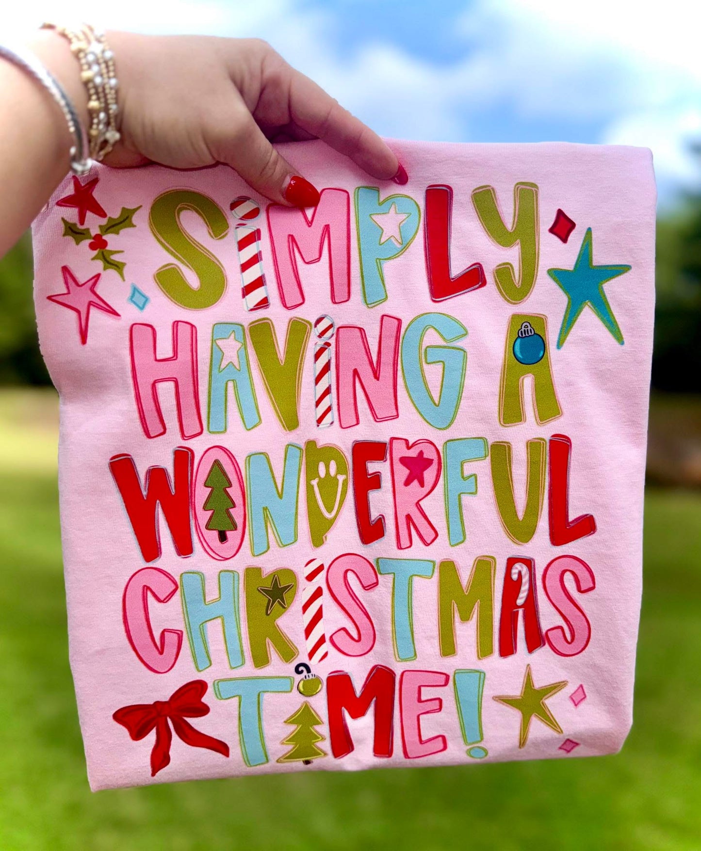 Pink tshirt with colorful text 'Simply Having a Wonderful Christmas Time!' held outdoors.