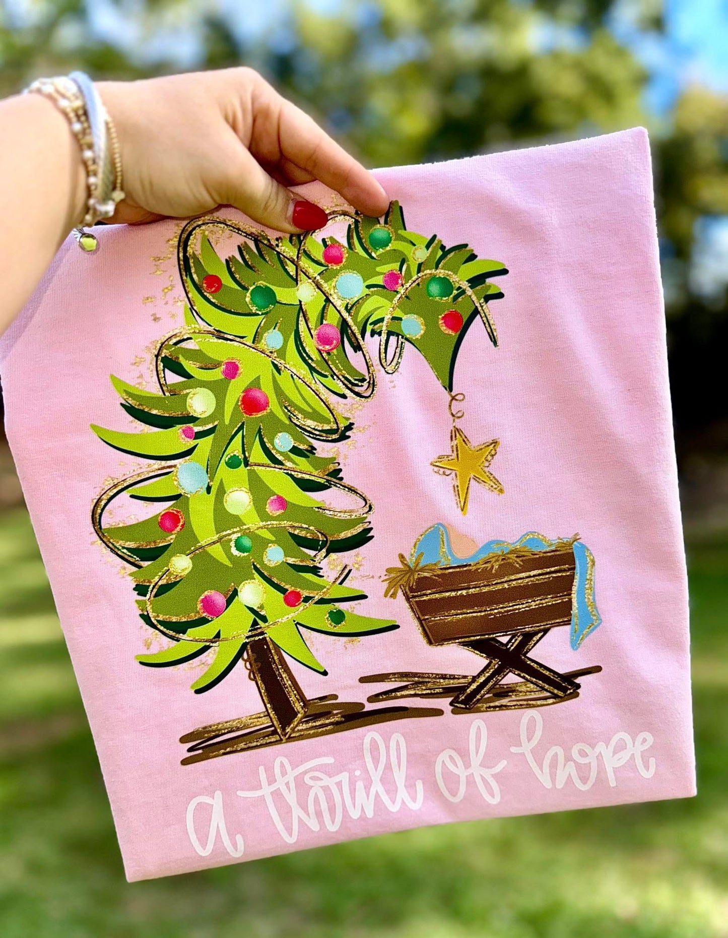 Pink sweatshirt with Christmas tree and manger design, held by a hand outdoors.
