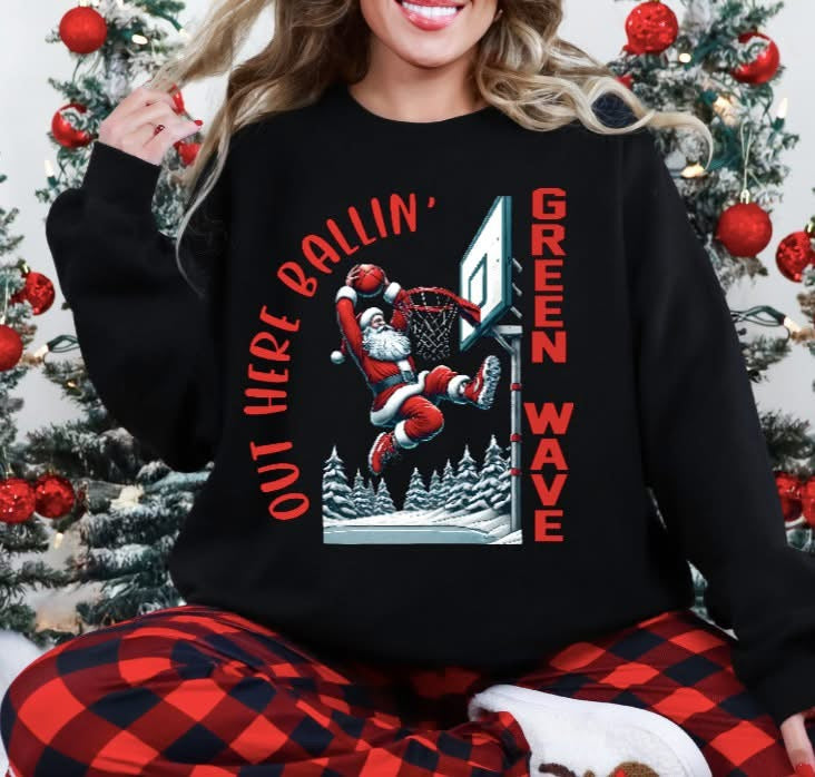 Person wearing a black sweatshirt with a graphic design in front of Christmas trees. Graphic is Santa dunking a basketball with wording saying "out here Ballin'" and "Green Wave" for the local school team 