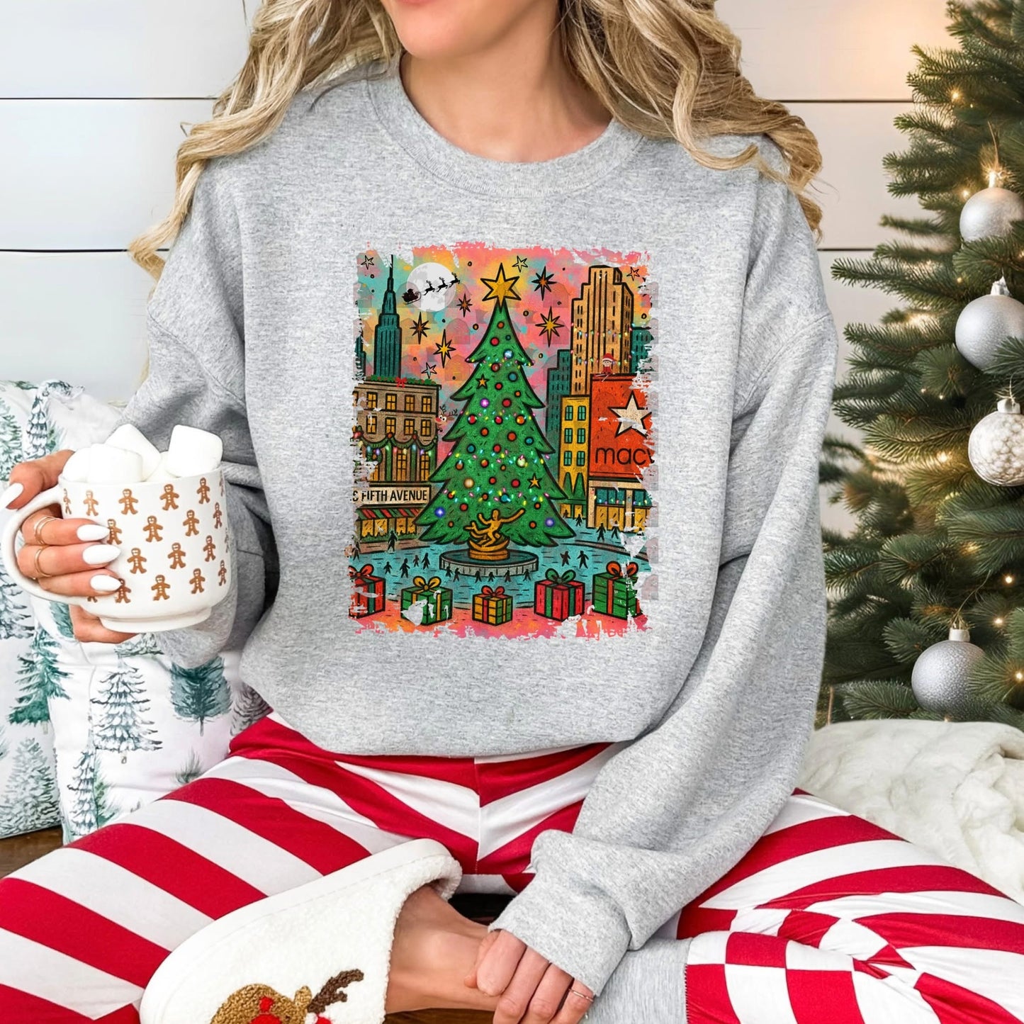Person wearing a festive sweater with a city Christmas-themed design, holding a mug, sitting in front of a decorated Christmas tree.
