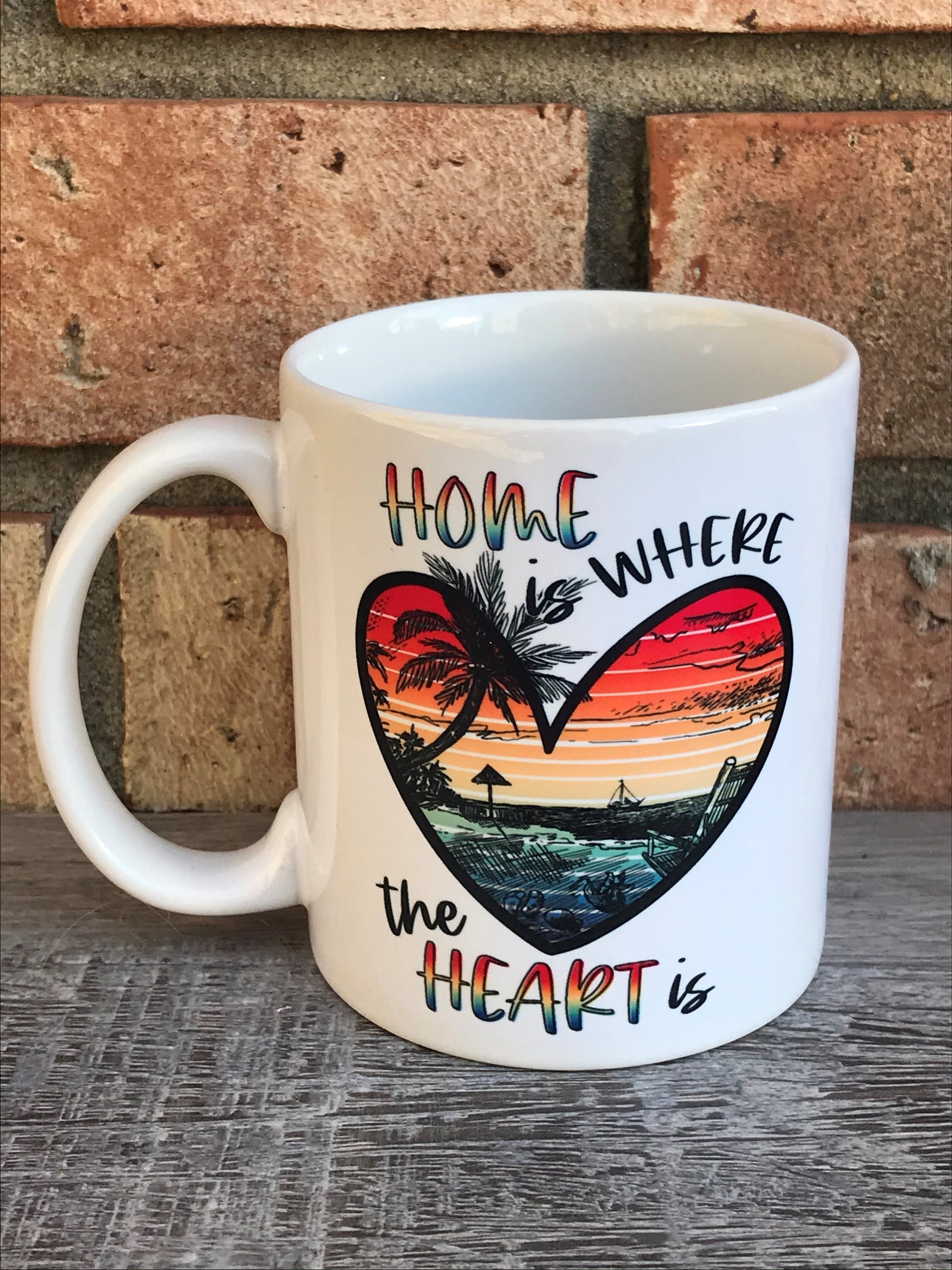 A white ceramic mug with a colorful beach-themed design, including palm trees and a sunset, with the text 'HOME IS WHERE THE HEART IS'