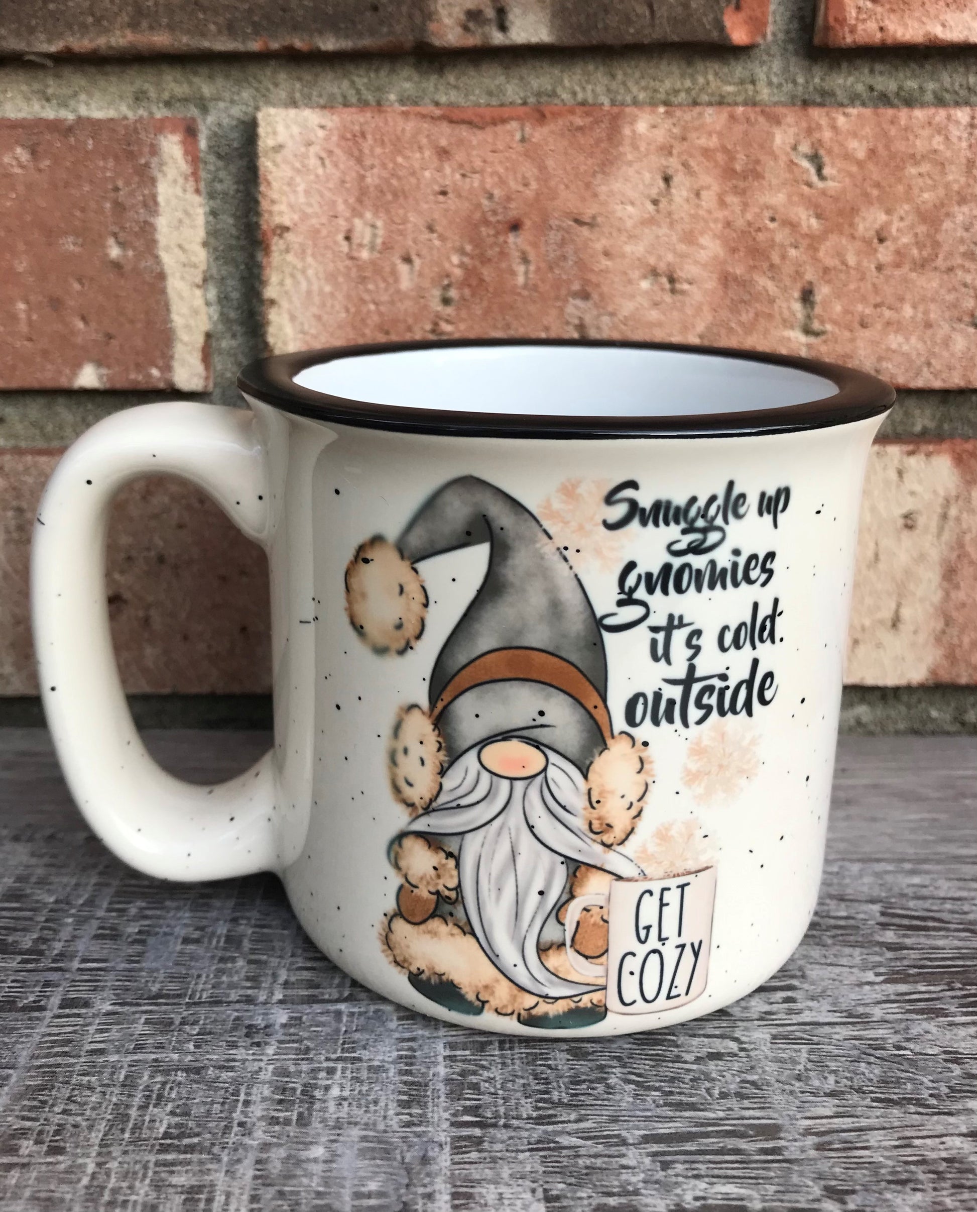 Camp style beige mug with speckled pattern that has a gnome on the front with a coffee cup that reads Snuggle up gnomies it’s cold outside 