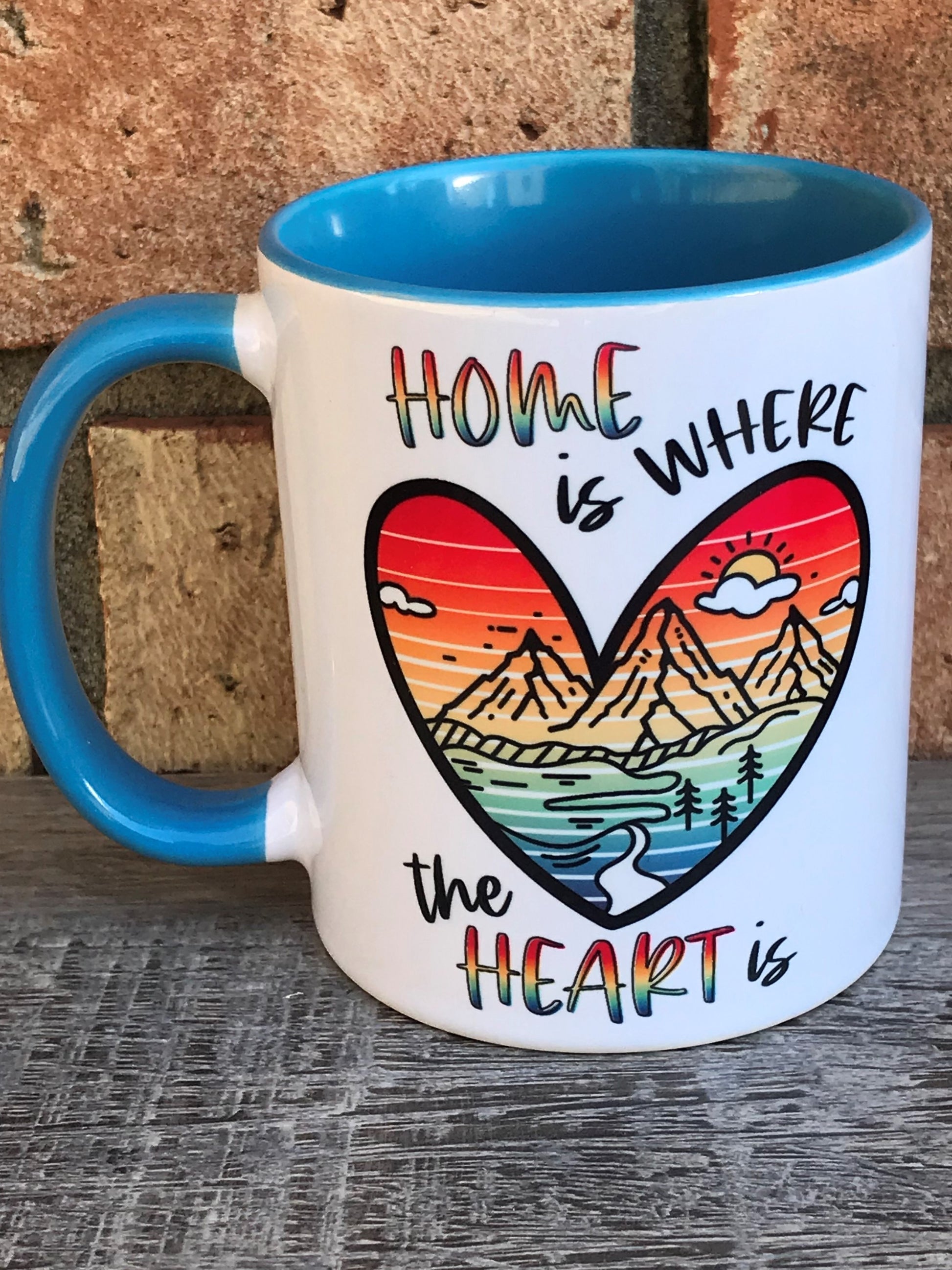 A white ceramic mug with blue interior and a mountain in a heart scene on the front that reads home is where The heart is 