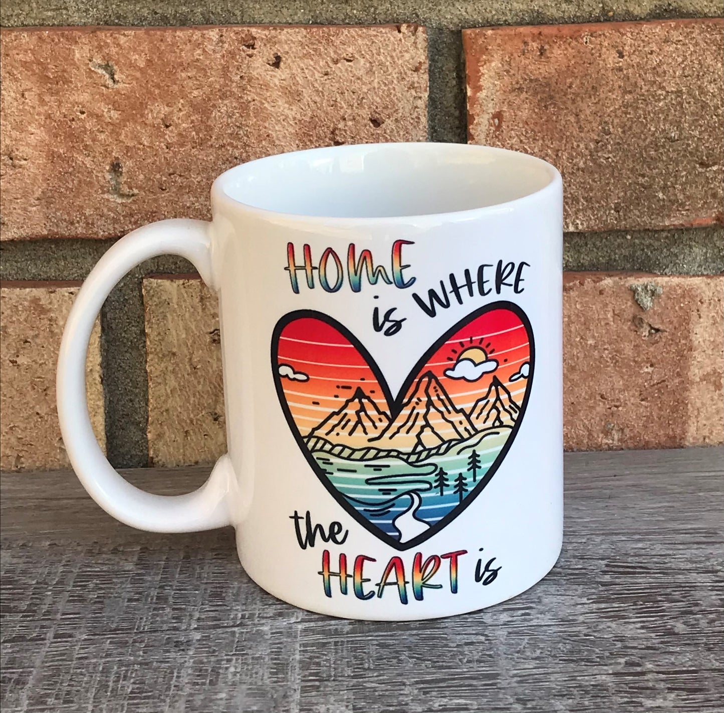 A white ceramic mug with a colorful mountain and heart design, and the text 'HOME IS WHERE THE HEART IS'.